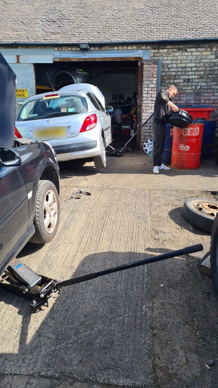 A mechanic changing a wheel in the yard beside the workshop, with a trolley jack in the foreground