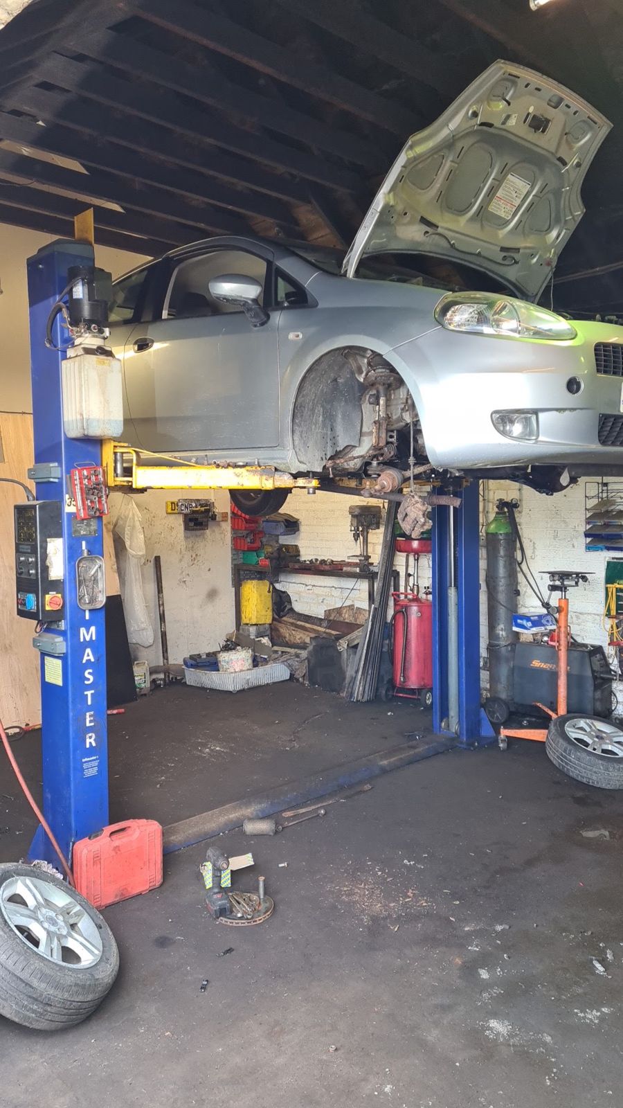 A silver hatchback up on the two-post lift with its front wheel off and bonnet open